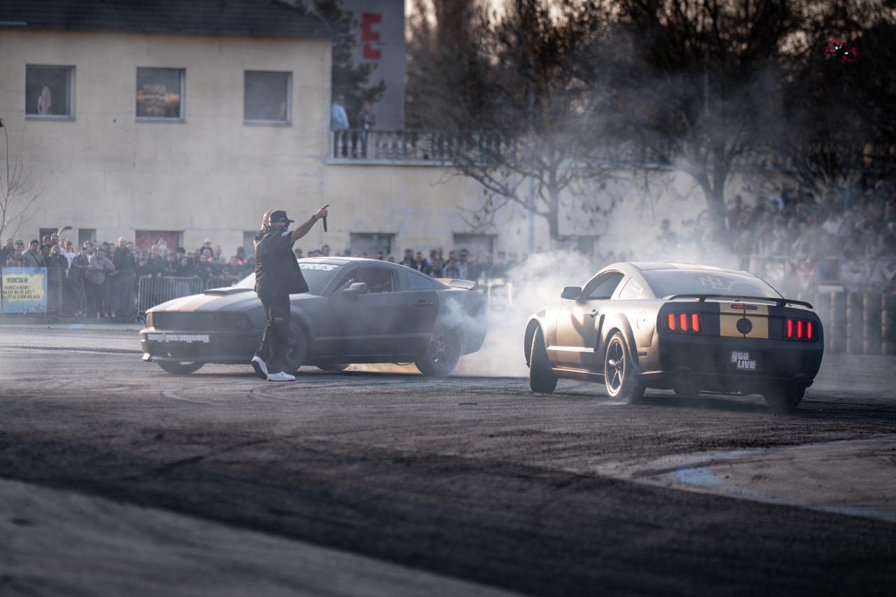 MC host standing in front of two matte black Ford Mustangs doing burnouts at GTA Live outdoor show