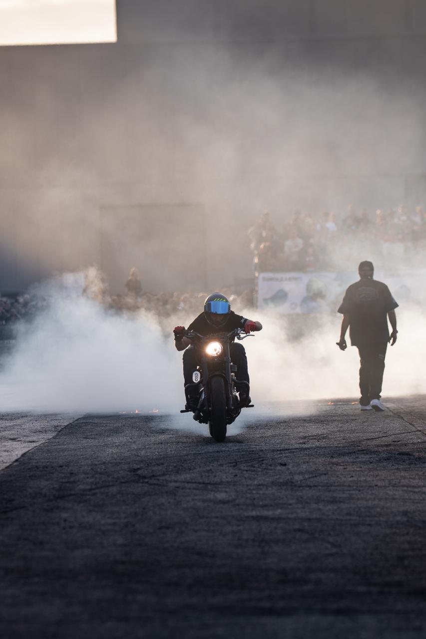 Motorcycle rider approaching head-on through white smoke cloud at outdoor stunt event