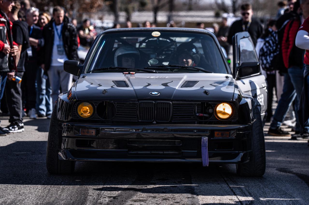 Black BMW E30 with flame hood graphics and yellow headlights facing crowd at outdoor car show