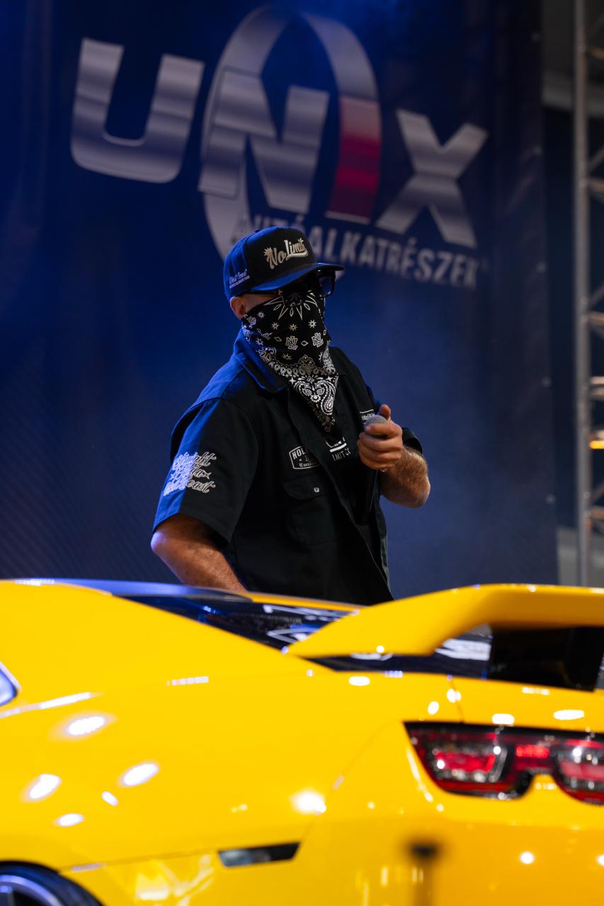 MC host in bandana and No Limit cap stands in front of yellow Camaro on UNX Alkatrészek stage
