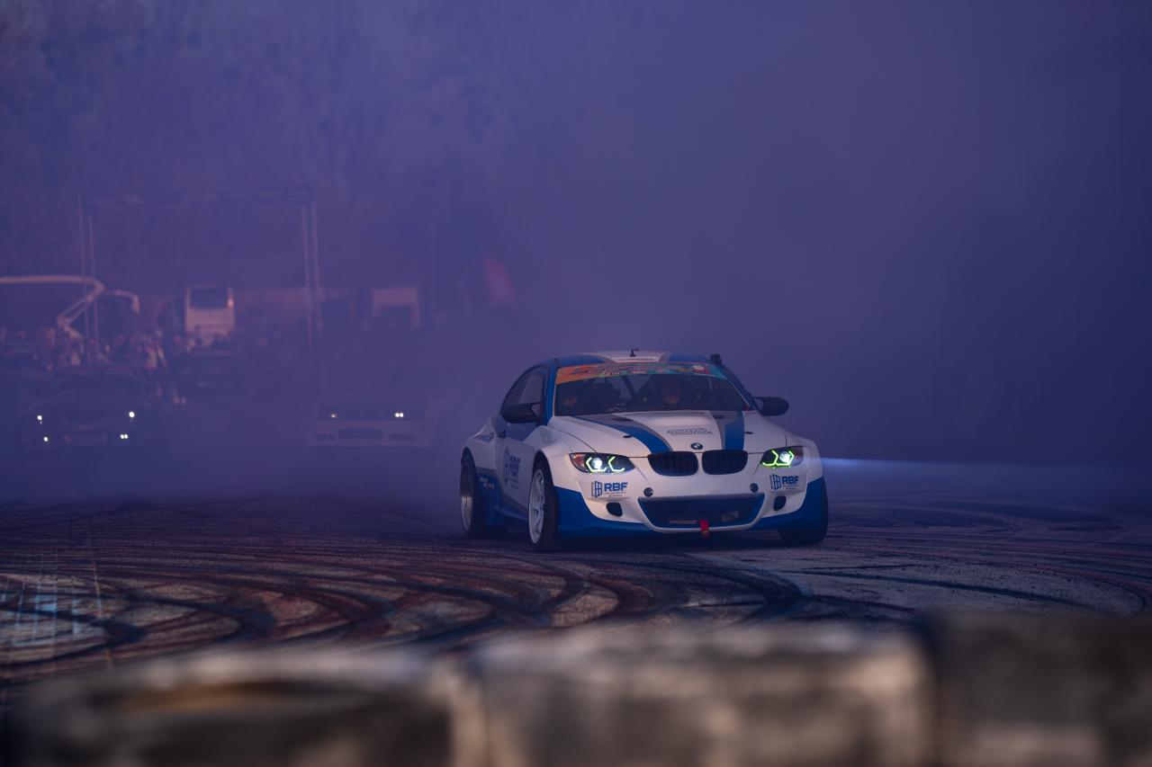 White and blue BMW RBF drift car sliding through fog and smoke at dusk motorsport event