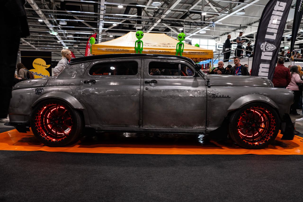 Custom vintage rat rod car with red LED wheels and alien figure sculptures on roof at indoor auto show