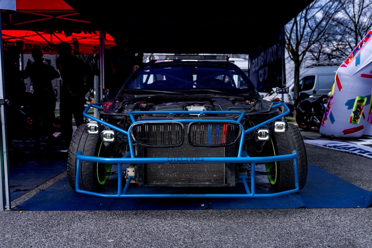 BMW race car with exposed blue roll cage and engine in outdoor event paddock