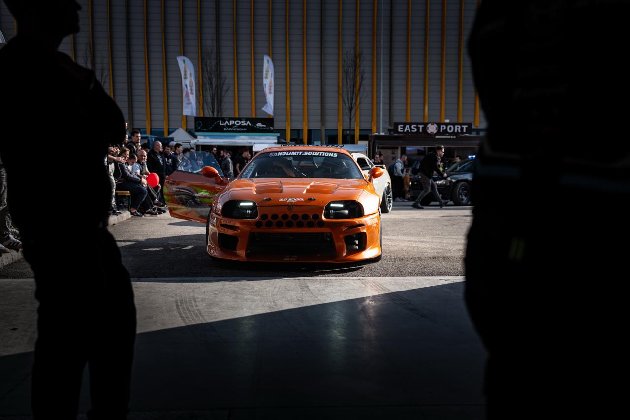 Orange Toyota Supra head-on at car show, framed through silhouetted spectators
